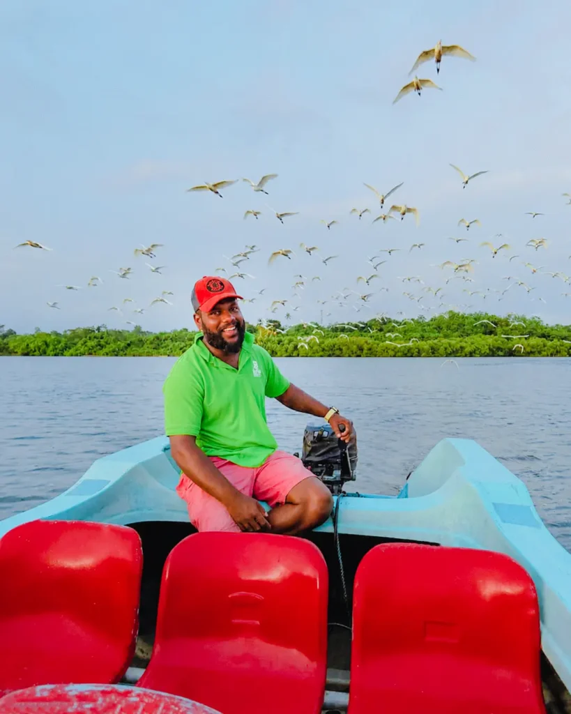 Smiling boat operator with herons flying above the Madu River wetlands